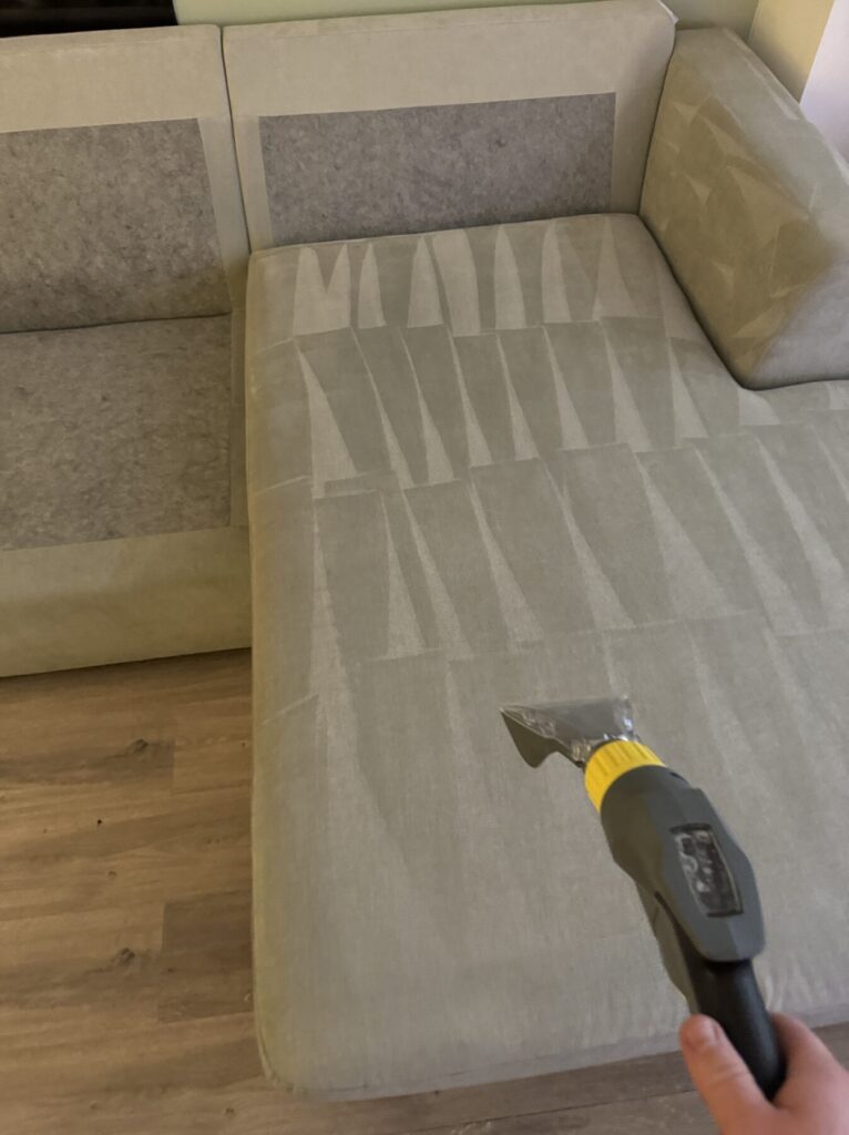 Professional upholstery cleaning tool extracting dirt and stains from a light grey fabric chaise lounge seat.